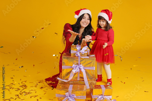 a mother in a red dress wearing a Santa Claus hat is sitting on a yellow background, a little daughter is standing next to her, gift boxes are standing next to them, confetti falls on them, a New Year