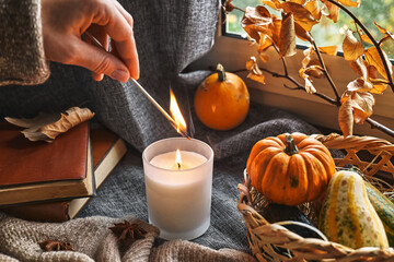 Hand with burning match lighting a candle on the windowsill with cozy autumn still life with pumpkins, knitted woolen sweater and books. Autumn home decor. Cozy fall mood. Thanksgiving. Halloween.