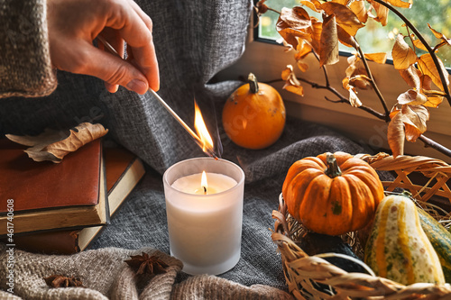 Fototapeta Naklejka Na Ścianę i Meble -  Hand with burning match lighting a candle on the windowsill with cozy autumn still life with pumpkins, knitted woolen sweater and books. Autumn home decor. Cozy fall mood. Thanksgiving. Halloween.