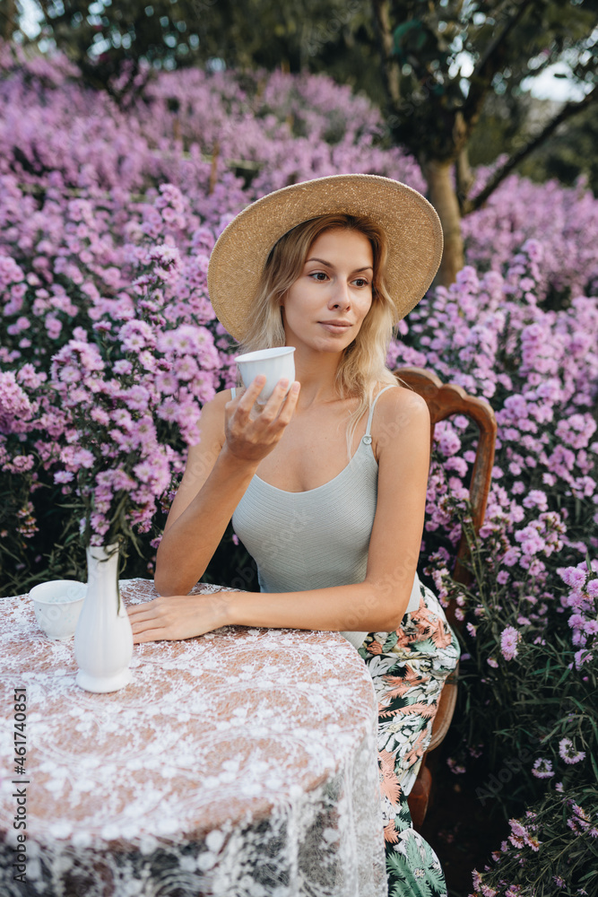 Fototapeta premium Young blonde woman sitting at the table with cup of tea in flower garden