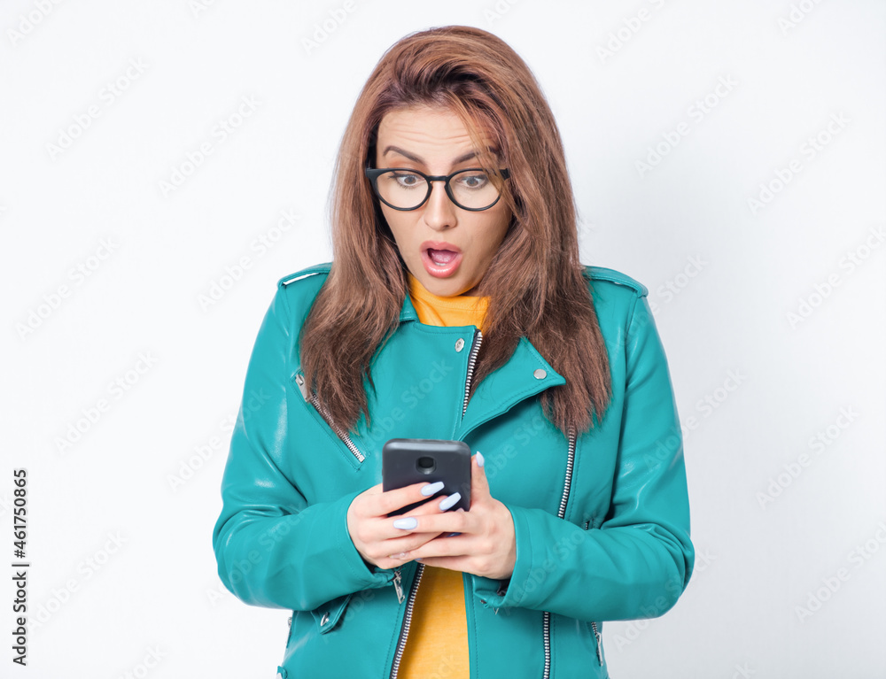 Unexpected message on smart phone. Surprised young woman with phone browsing, wearing green leather jacket, isolated on white background. Unbelievable