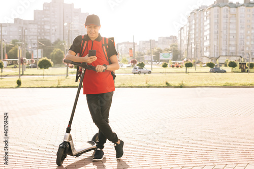 Asian man courier food delivery standing with electric scooter and thermal backpack uses smartphone confirm online customer client order. Deliveryman worker employee in red uniform.