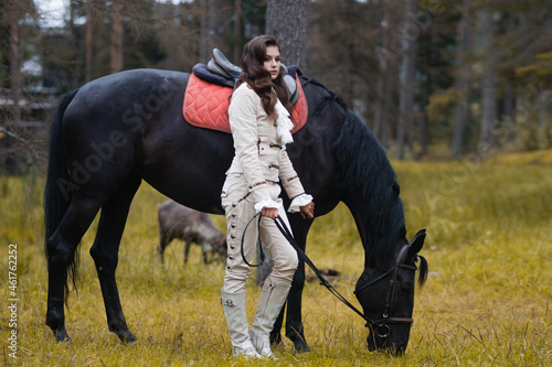 A young beautiful brunette rider next to a black mare in full ammunition, © Ulia Koltyrina