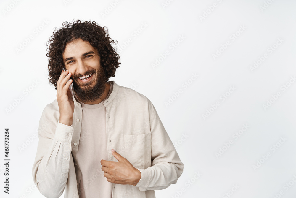 Handsome young man laughing and smiling during phone call, having conversation, standing over white background Handsome young man laughing and smiling during phone call, having conversation, standing over white background