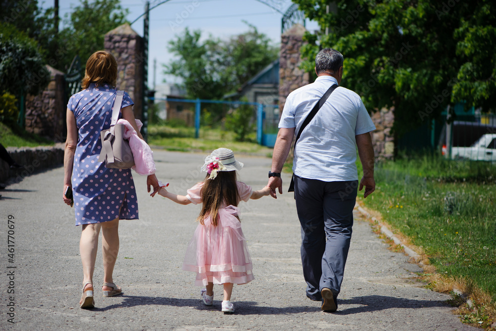 Obraz premium family walking in the park. mom dad and little girl walking holding hands. back view. daughter in a pink dress. friendly loving family concept, love. outdoor, walk in the fresh air, threesome