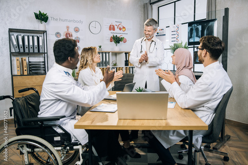 Team of multiracial medical specialists smiling and applauding after successful meeting at office room. Competent doctors sitting at table with various documents and modern gadgets.