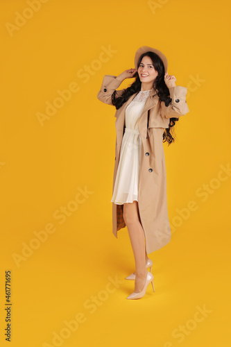 a young beautiful brunette girl with long hair in a white dress and a beige coat holds her beige hat yellow background