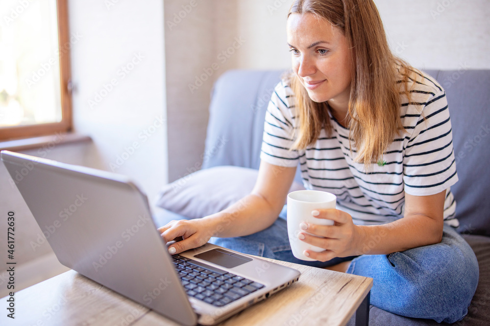 Beautiful young woman holding cup and laughing while looking at her laptop computer while sitting on the sofa near bright sunny window Beautiful young woman holding cup and laughing while looking at her laptop computer while sitting on the sofa near bright sunny window