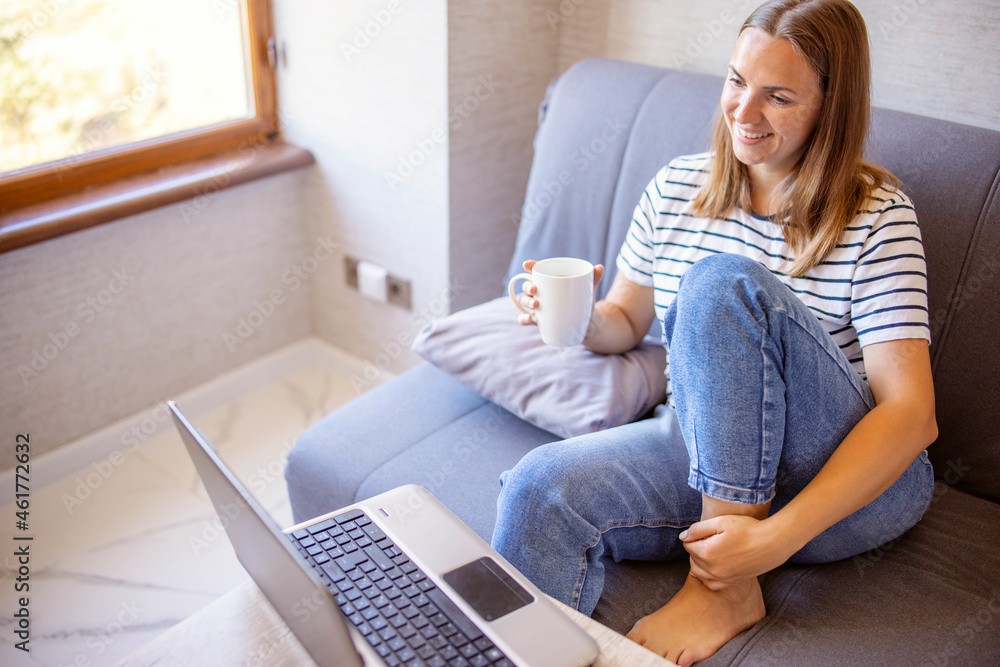 Beautiful young woman holding cup and laughing while looking at her laptop computer while sitting on the sofa near bright sunny window Beautiful young woman holding cup and laughing while looking at her laptop computer while sitting on the sofa near bright sunny window