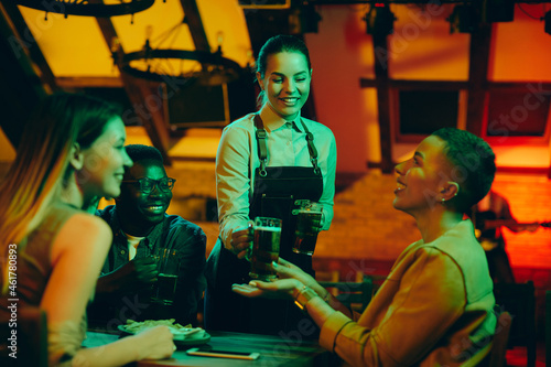 Young happy waitress serves beer to group of friends in pub.