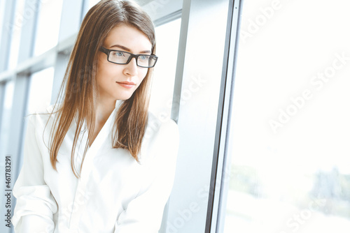 Beautiful female specialist with dark hair standing in modern office and smiling charmingly. Working on design, data analysis, plan strategy. Business people concept