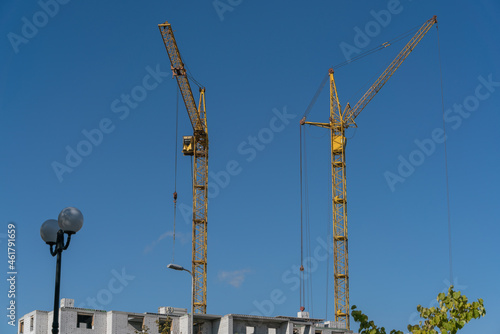 Two yellow construction cranes are working on building a house against a blue sky on a sunny day