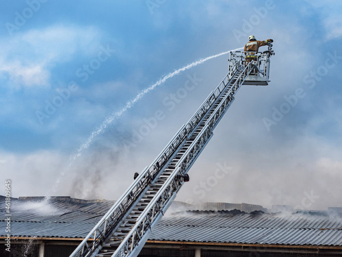 Firefighter on mechanized tower. Fire extinguishing with use of special equipment. Fire truck telescopic tower. Firefighter pours water on roof of building. Firefighter on tower on background of sky