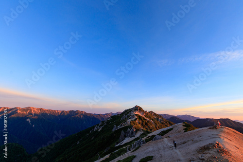 早朝に山頂へ向かう登山者と朝日に照らされた燕岳と山脈 © yuuki