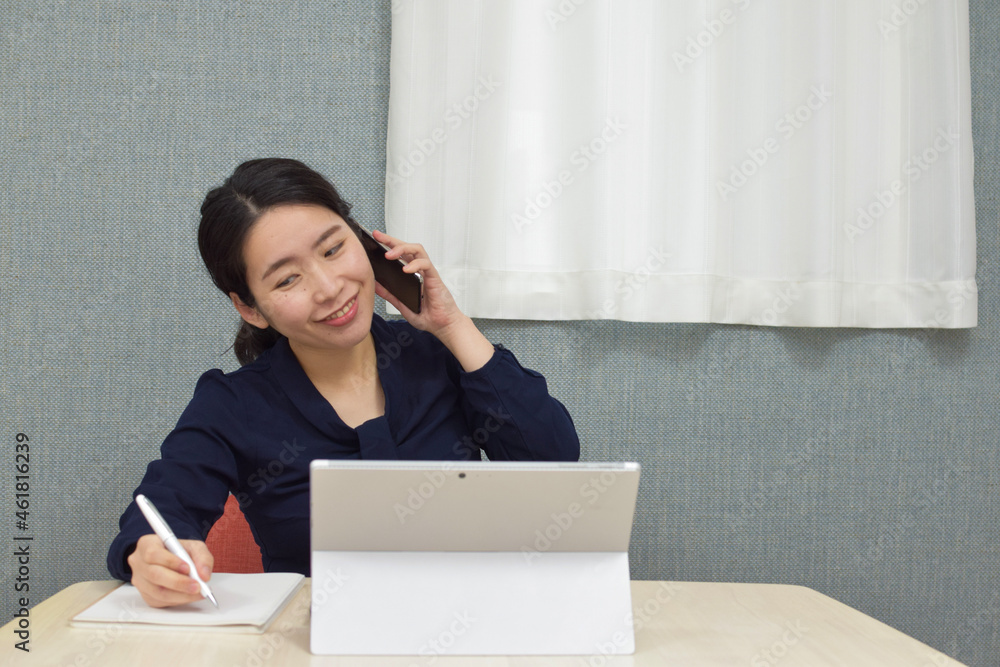 A young Asian woman working at home as telework talks on a telephone with smile and takes a memo on her notebook A young Asian woman working at home as telework talks on a telephone with smile and takes a memo on her notebook