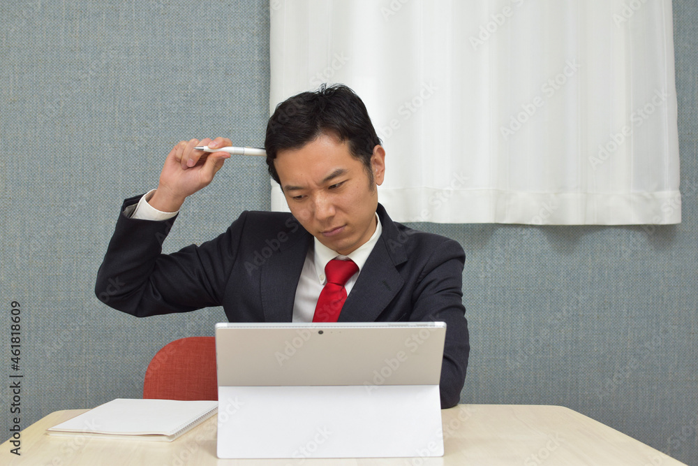 A young Asian man wearing business suit thinks about new idea and writes some memos on his notebook during telework at home A young Asian man wearing business suit thinks about new idea and writes some memos on his notebook during telework at home