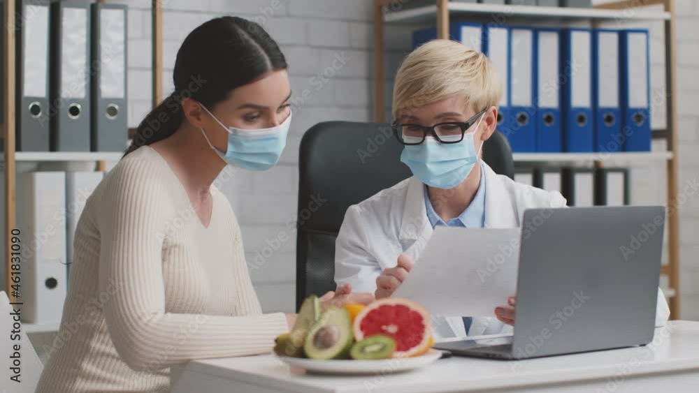 Young woman discussing dieting plan with professional doctor nutritionist, sitting in protective face masks in office Young woman discussing dieting plan with professional doctor nutritionist, sitting in protective face masks in office