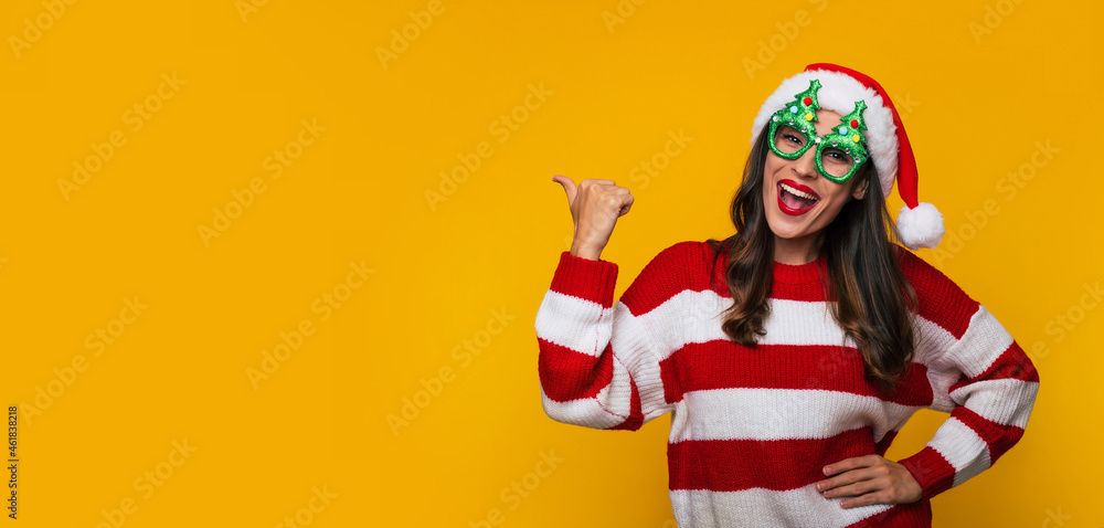 Banner photo of an excited beautiful modern woman in funny Christmas glasses and Santa hat is having fun while posing against a yellow wall