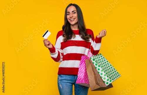 Close up photo of beautiful young happy and excited woman in a striped sweater with many colorful shopping bags and credit card in hands isolated on yellow background