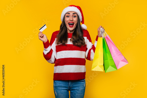 Happy stylish beautiful woman in Christmas hat with colorful shopping bags isolated on yellow background