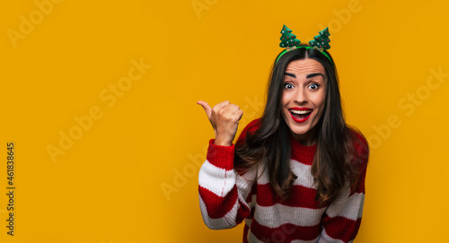Happy attractive funny young woman in Christmas tree hat on her head while she is pointing away isolated on a yellow background and having fun