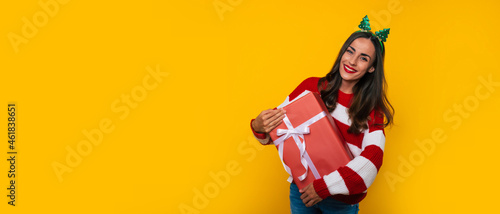 Wide banner photo of beautiful excited smiling woman with Christmas gift box in hands is having fun while posing on yellow background