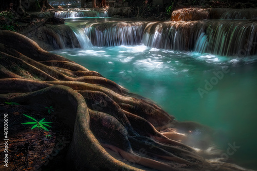 Erawan Waterfall at Kanchanaburi in Thailand during rainy season . A lot of water flows down through the layers of limestone with a large root of tree as foreground.