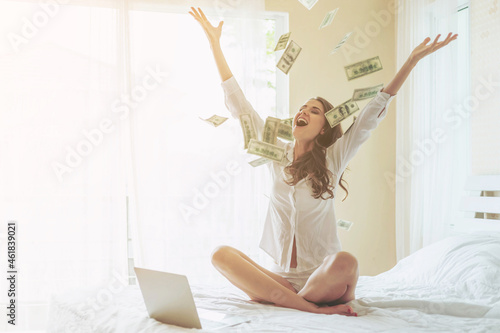 Beautiful woman sitting on the bed in front of her laptop , sprinkle money on the bed with a happy smile  Because she profits from investing in online stocks - start up online business concept photo