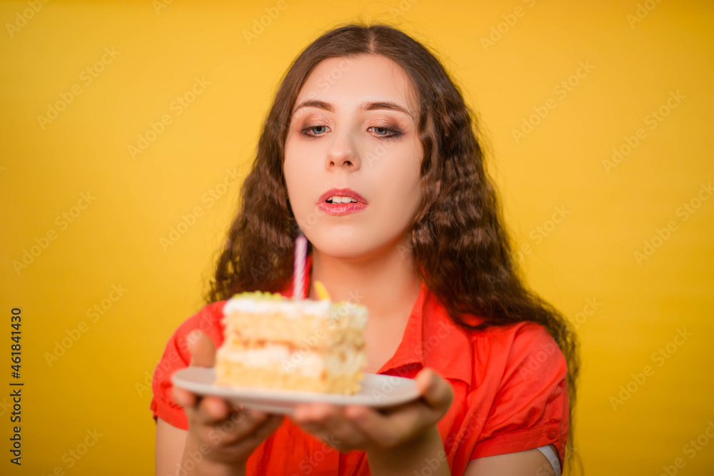 Portrait of a girl in a red shirt with a piece of cake and candle on a white plate in her hands isolated on a yellow background.