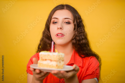 Portrait of a girl in a red shirt with a piece of cake and candle on a white plate in her hands isolated on a yellow background.