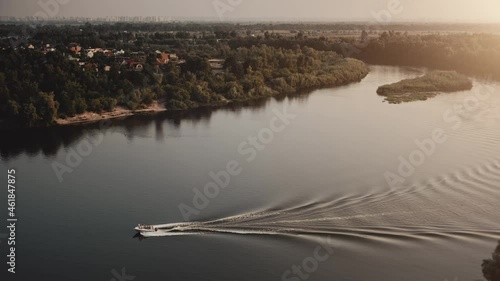 Modern motor boat rides in river countryside landscape. White yacht ship with people having fun, enjoy river voyage in rural city. Sunset over marine village. Summer vacation, recreation. Drone flight
