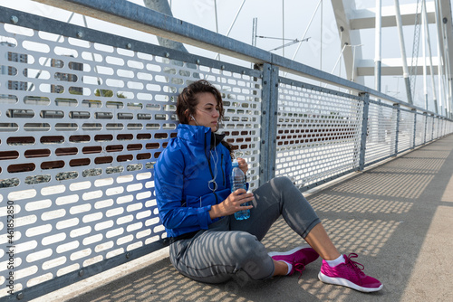 Young beautiful fitness sporty jogger woman squats as taking a break on the bridge drinking water for hydration after cardio workout and training for healthy life by running outdoors