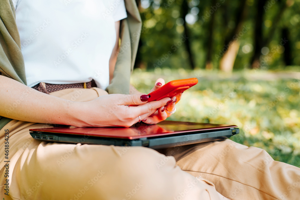 Close-up of young woman holding mobile phone, chatting, browsing social media, sending messages outdoors. Selective focus of female hands using red smartphone in nature. Cropped image