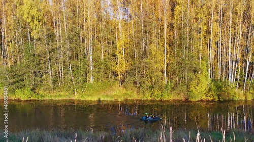 a boat is sailing on a calm river in an autumn forest and two people are rowing with oars

