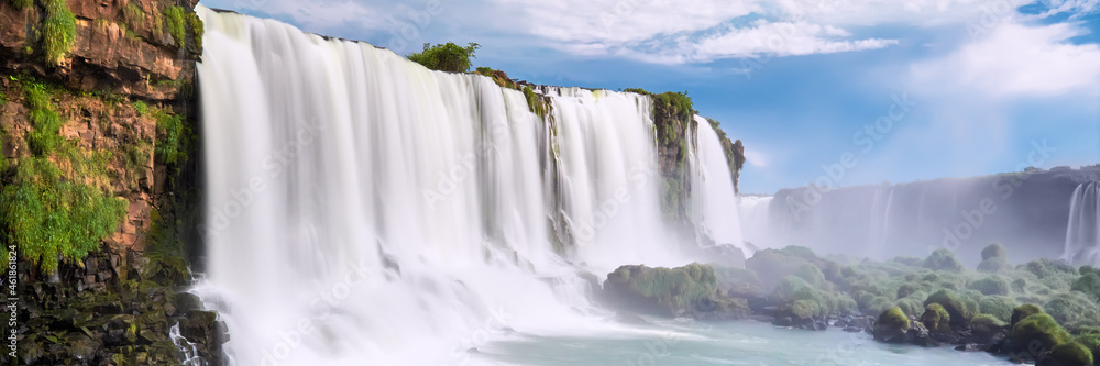 Naklejka premium Iguazu waterfalls in Argentina, view from Devil's Mouth. Panoramic view of many majestic powerful water cascades with mist. Panoramic image with blue sky and clouds.