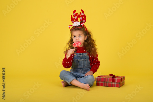 Little girl for the Christmas holidays with a gift and a lollipop on a yellow background to copy space