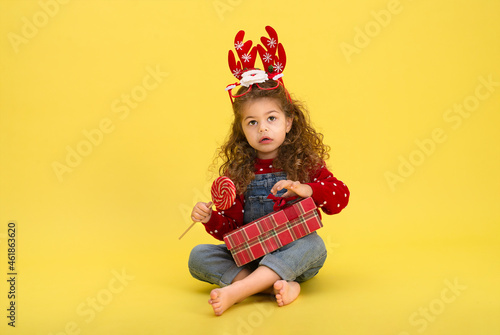 Little girl for the Christmas holidays with a gift and a lollipop on a yellow background to copy space