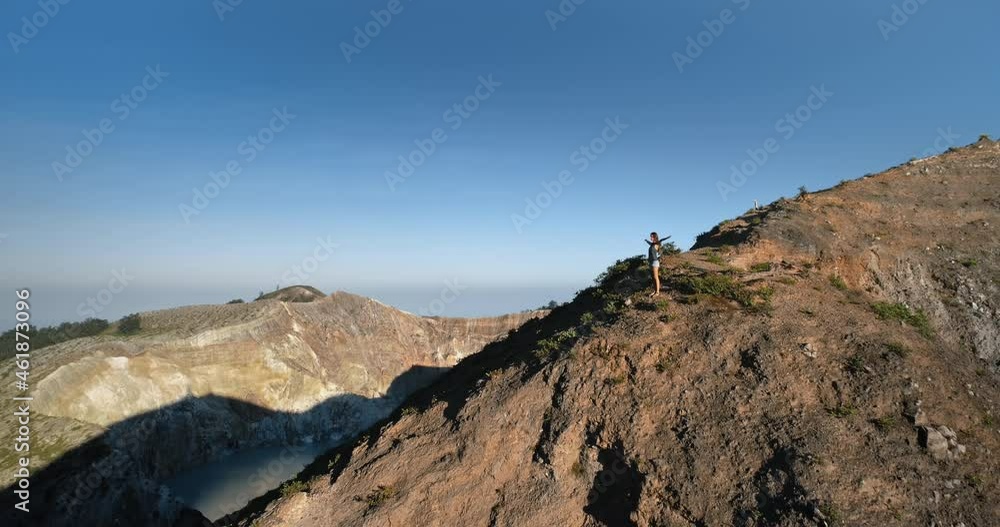Woman standing on mountain top with hands rise up. Volcano crater lake, blue sky in background. Beautiful wild nature landscape. Exotic summer vacation, travel, outdoor tourism. Cinematic drone flight Woman standing on mountain top with hands rise up. Volcano crater lake, blue sky in background. Beautiful wild nature landscape. Exotic summer vacation, travel, outdoor tourism. Cinematic drone flight