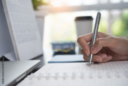 Close up of business woman hand with a pen writing notes on spiral notepad, planning work project, event schedule with calendar on office desk
