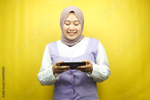 Beautiful asian young muslim woman with hands holding smartphone, playing game, smiling happily, victory, success, isolated on yellow background, advertising concept