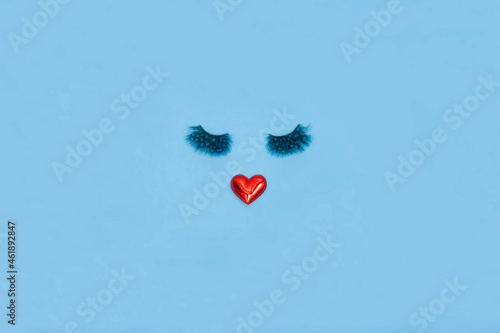 Creative woman face made of eyelashes and red heart lips on the blue background. Minimal beauty concept. Flat lay. Valentine makeup creative concept.