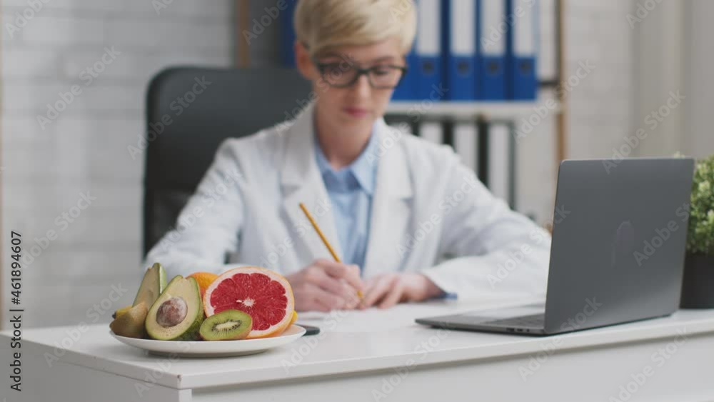 Zoom in shot of middle aged woman nutritionist working with papers at office, plate with fruit variety on table Zoom in shot of middle aged woman nutritionist working with papers at office, plate with fruit variety on table