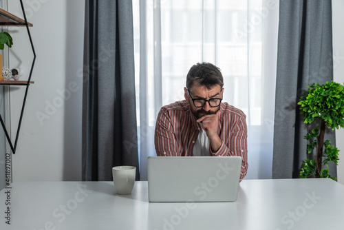 Thoughtful businessman in eyewear looking at computer screen, sitting at table at home. Pensive confused young man thinking of problem solution stuck with task, working with laptop remotely at home