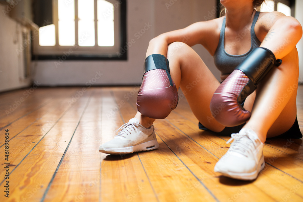 Close-up of young woman relaxing after training. Woman in modern sport clothes sitting on floor, white sneakers, red boxing gloves. Sport, healthy lifestyle, boxing concept Close-up of young woman relaxing after training. Woman in modern sport clothes sitting on floor, white sneakers, red boxing gloves. Sport, healthy lifestyle, boxing concept