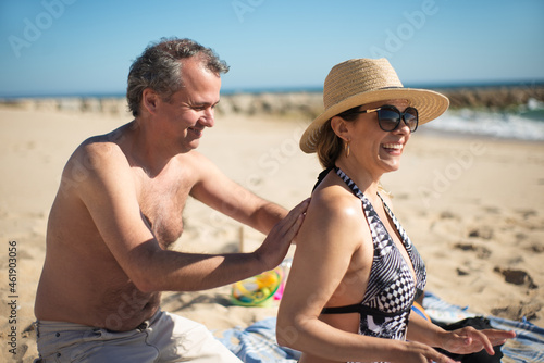 Happy wife and husband at beach. Mid adult man applying sunscreen on womans back. Family, vacation, outdoor activity concept