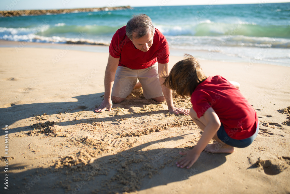 Happy father and son at sea coast. Mid adult man and schoolboy in red t-shirts drawing at sand. Family, vacation, outdoor activity concept Happy father and son at sea coast. Mid adult man and schoolboy in red t-shirts drawing at sand. Family, vacation, outdoor activity concept