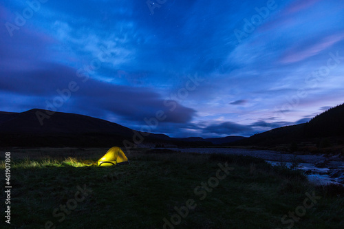 illuminated tent on meadow besides a river under a cloudy sky with star during blue hour  Scotland