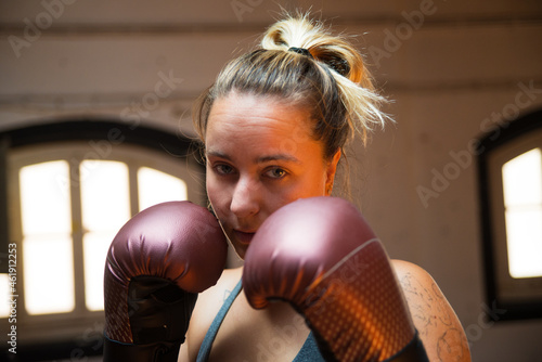 Portrait of young woman at boxing training session. Attractive woman in sport clothes standing at gym in combative position, looking at camera. Sport, healthy lifestyle, boxing concept