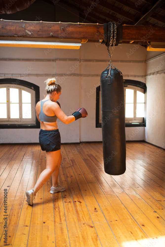 Back view of young woman at kickboxing training. Female boxer hitting huge punching bag at boxing studio. Sport, healthy lifestyle, boxing concept