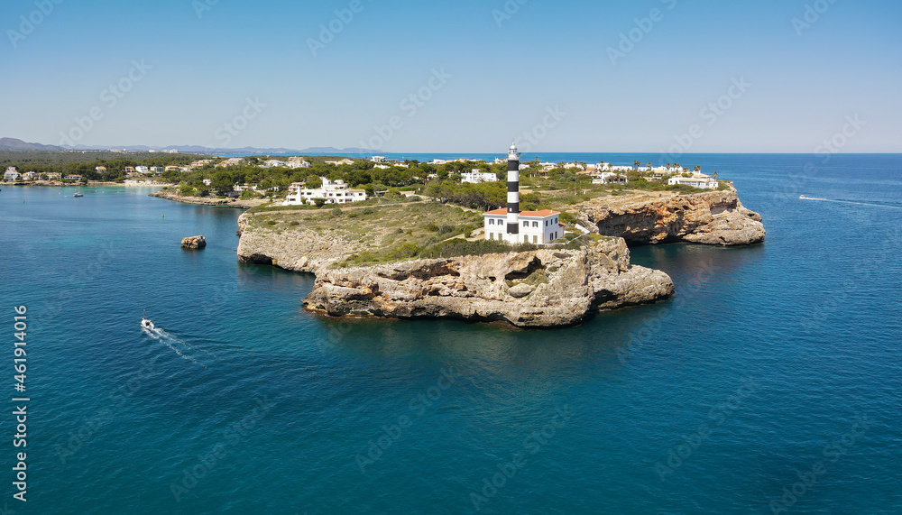 The lighthouse of Porto Colom on the Spanish Mediterranean island of Mallorca in sunshine in summer. A boat drives towards the harbor. In the foreground are the cliffs. The lighthouse of Porto Colom on the Spanish Mediterranean island of Mallorca in sunshine in summer. A boat drives towards the harbor. In the foreground are the cliffs.